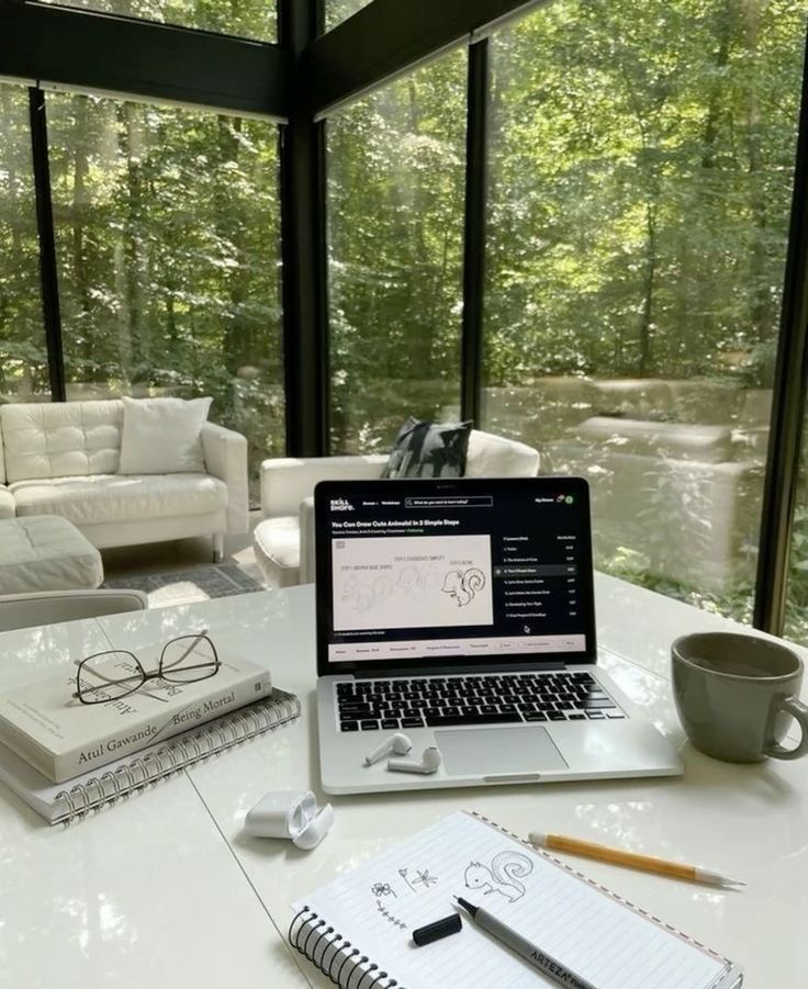 Minimalistic 2025 Daily Weekly Monthly Planner - Handy Home Home office with laptop and sketchpad in a glass-enclosed room, surrounded by lush greenery, creating a serene workspace atmosphere.