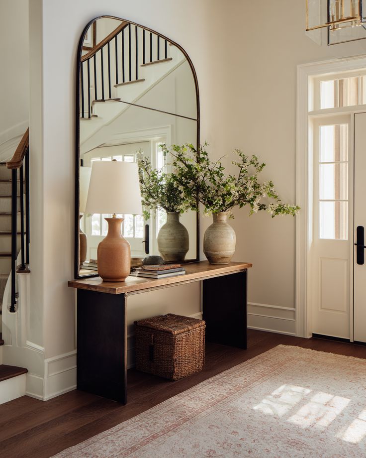 Cozy entryway with large mirror, elegant console table, vase with branches, lamp, basket, and staircase in background. Inviting decor.