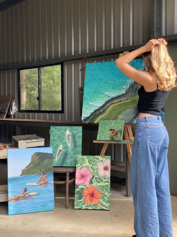 Artist in a studio with colorful ocean-inspired paintings on display, including surfers and floral elements, under natural light.