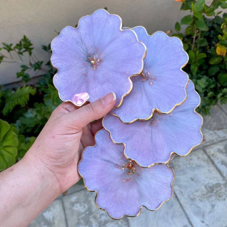 Hand holding four purple resin coasters with gold edges, shaped like flowers, against a garden background.