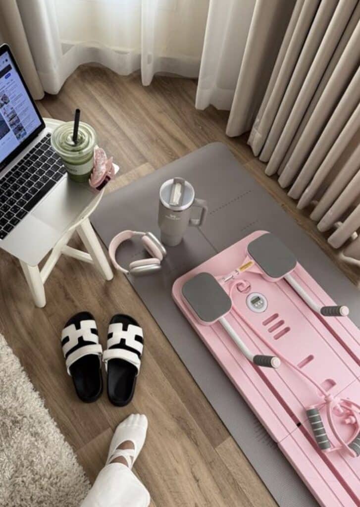 Cozy home workout setup with yoga mat, pink stepper, laptop, green smoothie, headphones, and slippers on wooden floor.