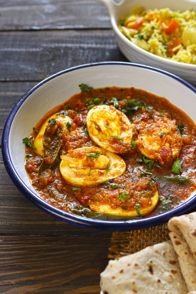 Punjabi Egg Curry Recipe - Handy Home Spicy egg curry in a bowl with rich tomato gravy, garnished with cilantro, accompanied by mixed vegetable rice and flatbreads.
