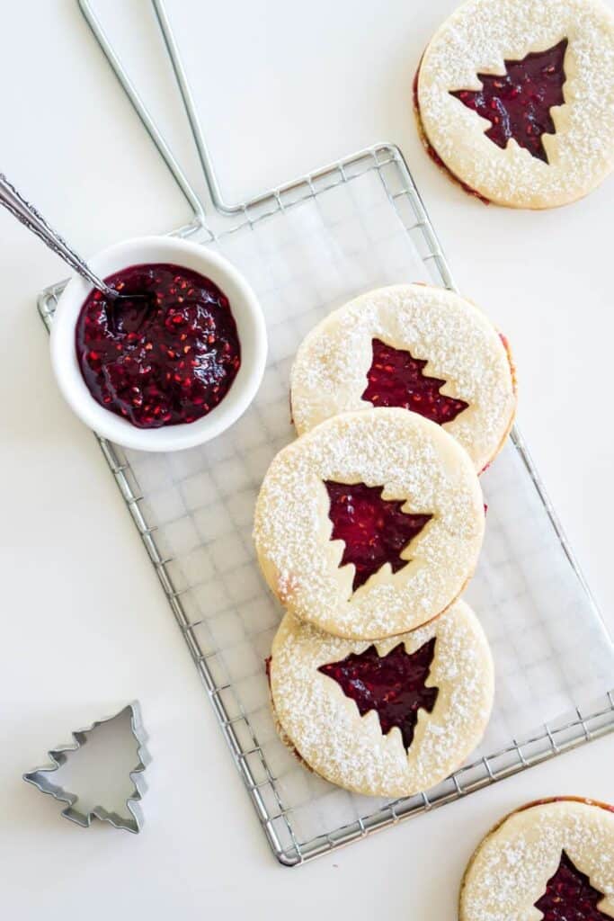 Raspberry Linzer Cookies - Handy Home Holiday cookies with raspberry jam filling featuring Christmas tree cutouts on a cooling rack. Perfect festive desserts.