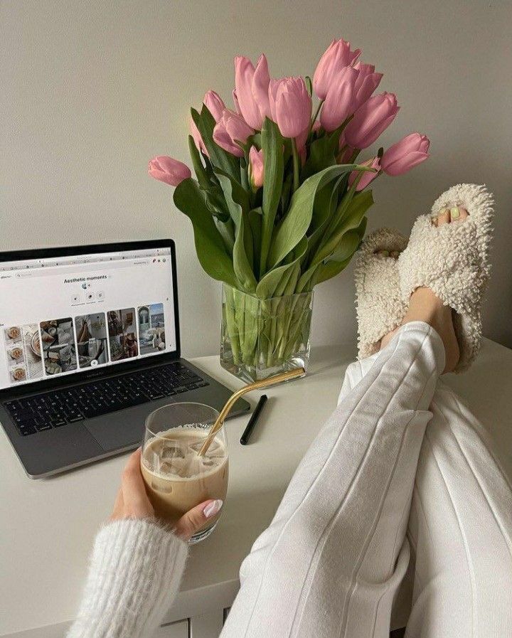 Cozy workspace with pink tulips, open laptop, and iced coffee on a white desk. Relaxed vibe with feet in fluffy slippers.