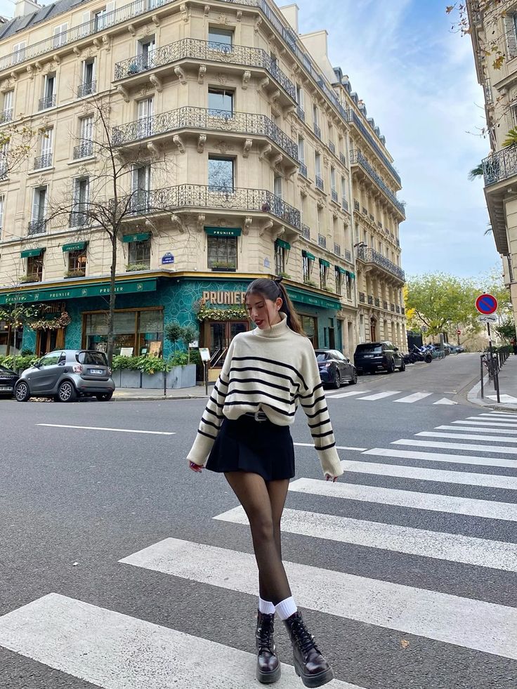 Young woman crossing a Parisian street wearing a striped sweater and skirt, stylish city scene with classic architecture in the background.