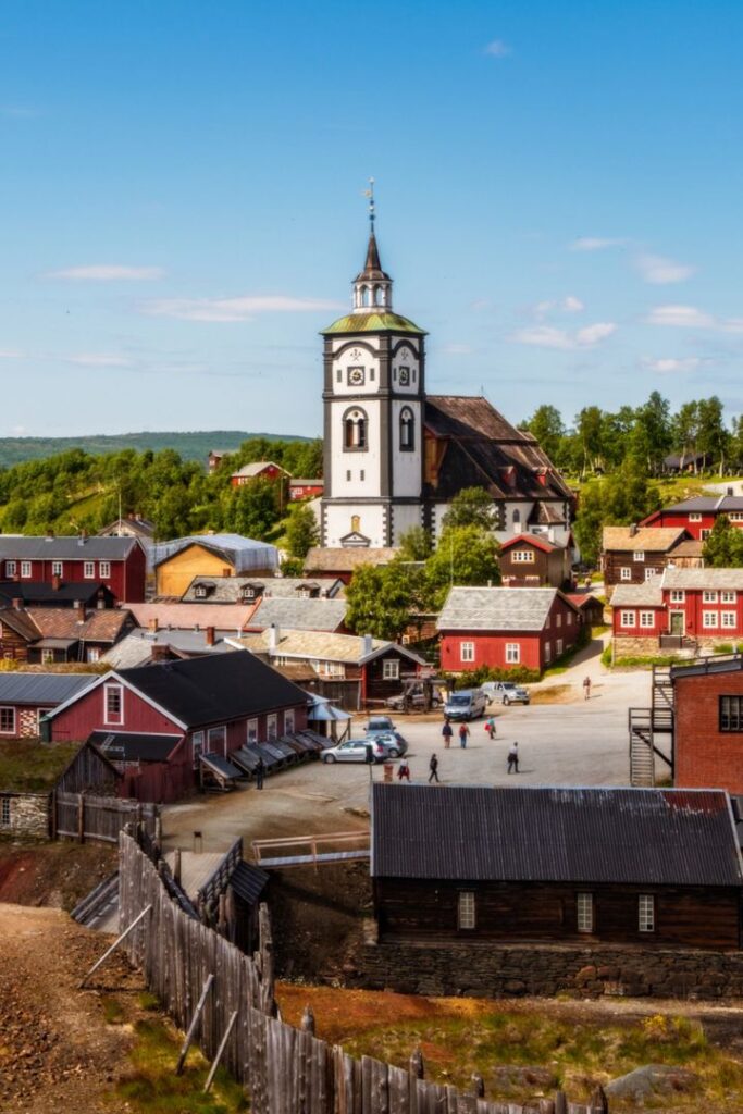 Røros Church Norway - Handy Home Historic Nordic town with a prominent church tower, surrounded by colorful wooden buildings and lush greenery under a blue sky.