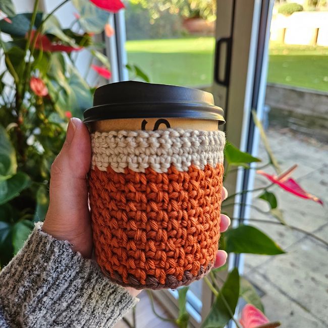 Hand holding a coffee cup with an orange crochet cozy, near indoor plants and glass door. Cozy autumn vibes.
