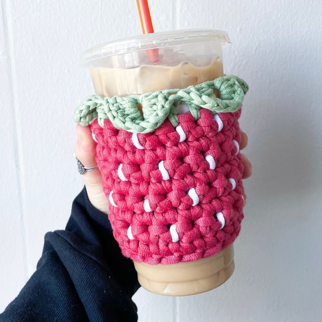 Hand holding iced coffee with a strawberry-themed crochet cozy, featuring a green leafy top and red berry pattern.