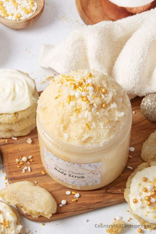 Sugar cookie body scrub in a jar with festive sprinkles, surrounded by cookies and a soft towel, on a wooden board.