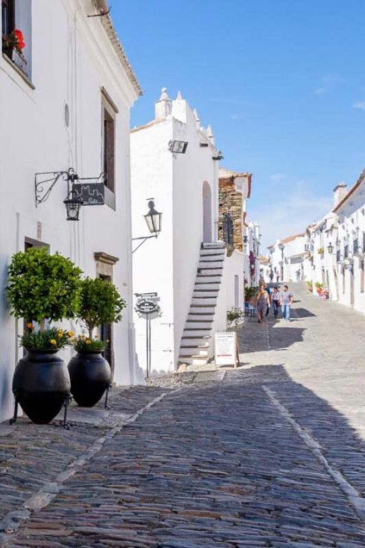 Tour Monsaraz - Handy Home Charming narrow street in a whitewashed village with cobblestone path, potted plants, and sunny blue sky. Ideal travel destination.