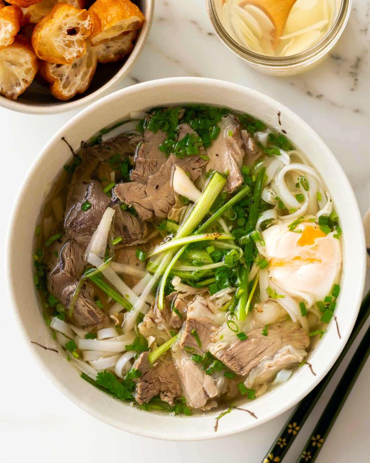 Vietnamese Recipes - Handy Home Bowl of Vietnamese pho with beef, rice noodles, herbs, and a poached egg, served with crispy breadsticks on a marble table.