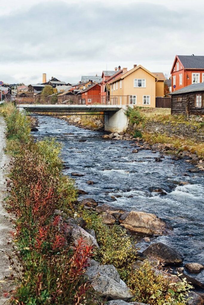 Visit Norway A Food Tour - Handy Home Scenic view of a charming river flowing through a quaint village with colorful houses and a bridge on a cloudy day.