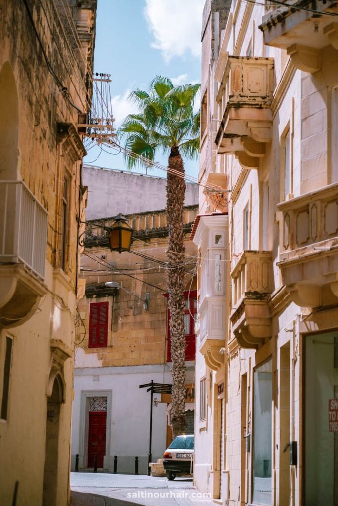 Wat te doen op Gozo_ 9 Leukste Bezienswaardigheden Malta - Handy Home Charming narrow street in sunlight, featuring a palm tree and traditional architecture with balconies.