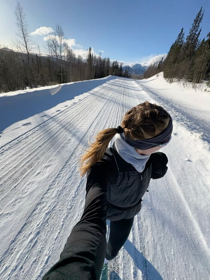 Woman jogging on a snowy mountain road, surrounded by trees and clear blue sky, enjoying a sunny winter day.
