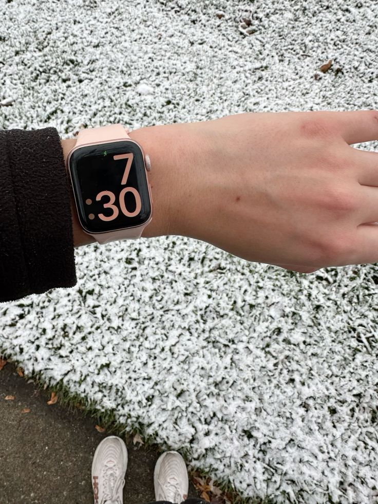 A wrist wearing a smartwatch displays 7:30 over a snowy lawn, with sneakers visible on the path.