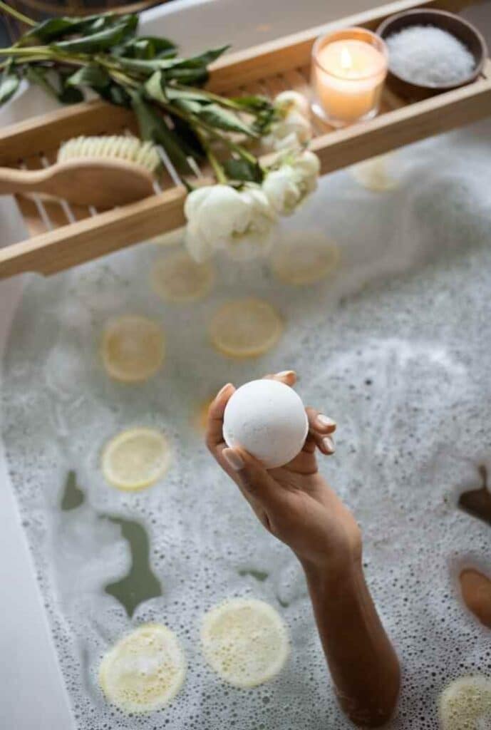 Hand holding a bath bomb above a relaxing bath with lemon slices, flowers, and candle for a soothing spa experience.