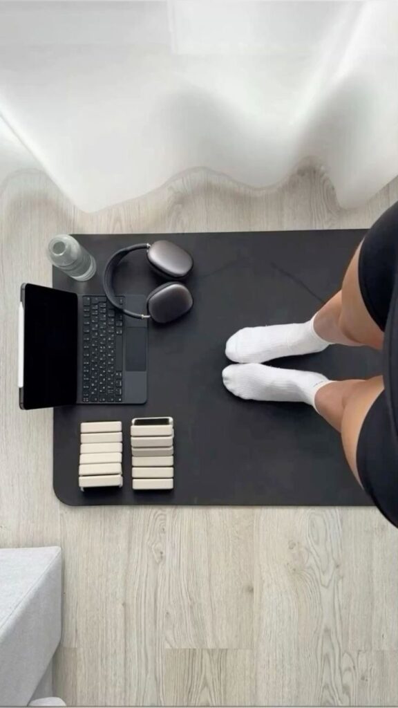 Person standing on black mat with headphones, laptop, water bottle, blocks, short sports shorts and socks. Minimalist home workout setup.