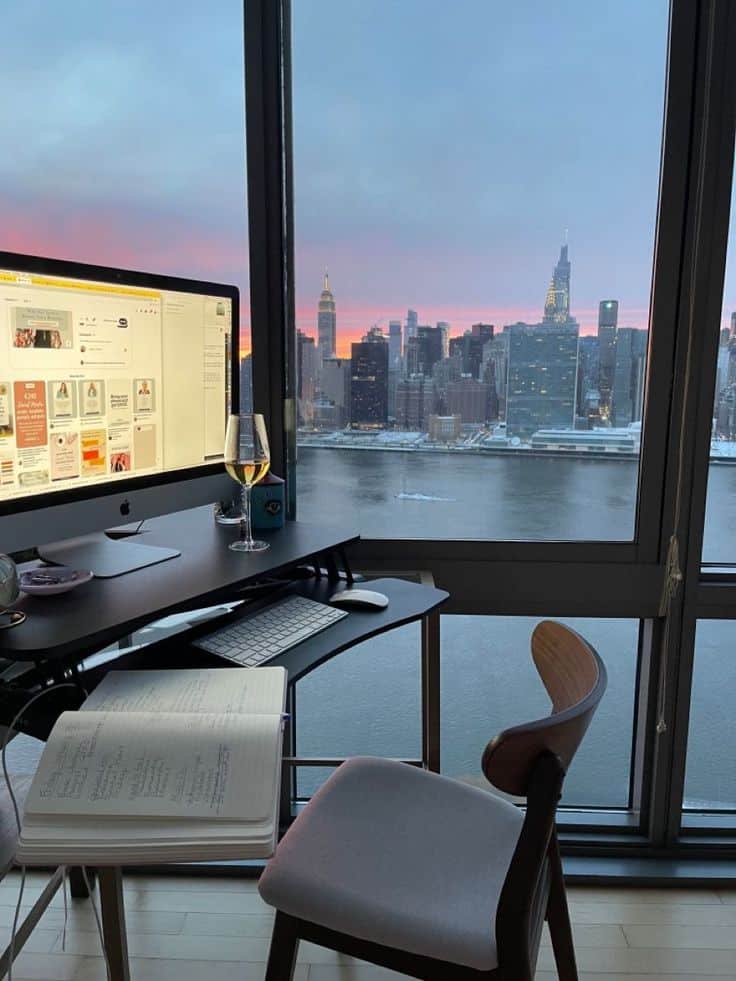 Home office setup with desktop computer, glass of wine, and notepad, overlooking a city skyline at sunset through large windows.