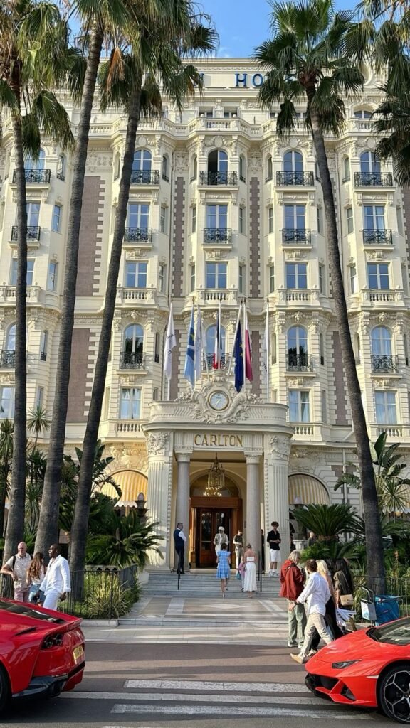 Grand hotel entrance with palm trees, luxury cars, and people arriving under flags. Elegant architecture and sunny setting.