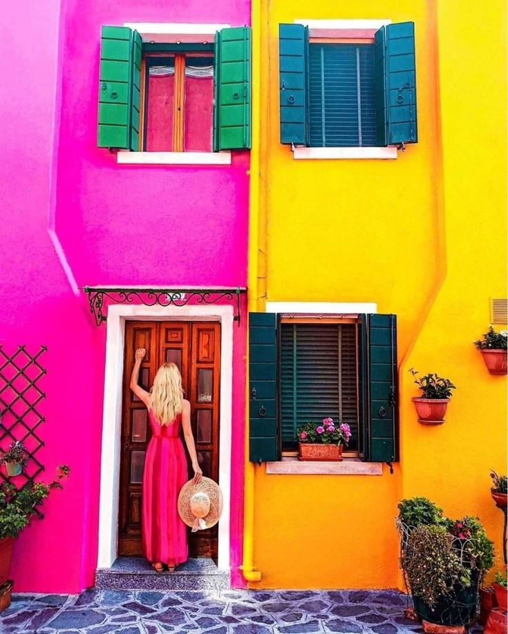 adfgve - Handy Home Woman in pink dress and hat at vibrant pink and yellow building with green shutters, colorful facade and potted plants.