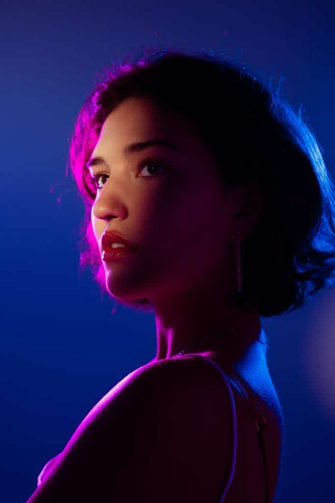 aegbetgb - Handy Home Portrait of a woman in vibrant pink and blue lighting, looking thoughtful and elegant with short hair and chandelier earrings.