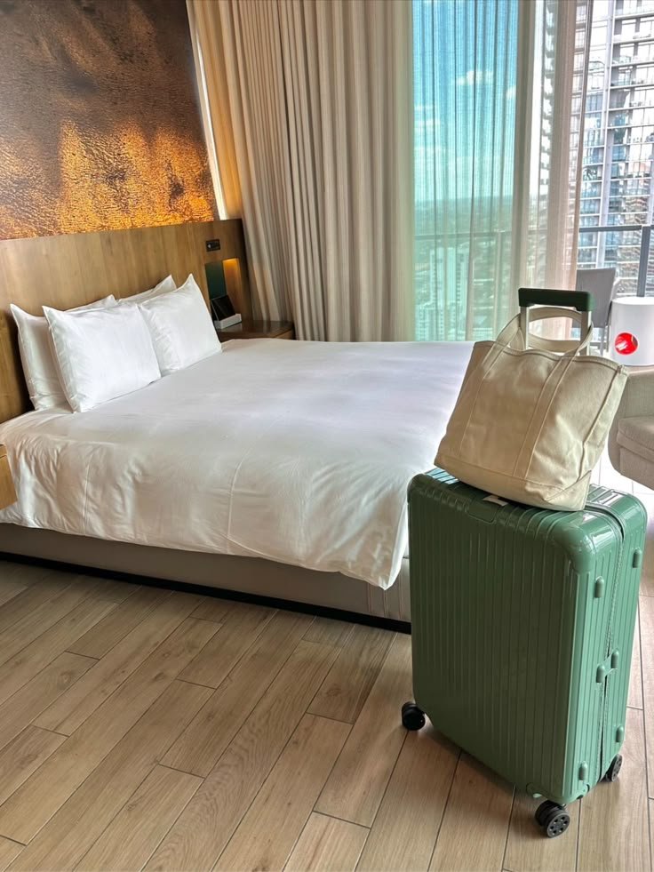 Modern hotel room with a cozy bed, green suitcase, and city view through large windows. Perfect for a comfortable stay.
