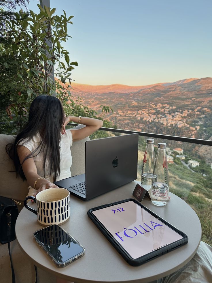 Woman enjoying a scenic mountain view while working on a laptop, with coffee, tablet, and water bottles on the table.