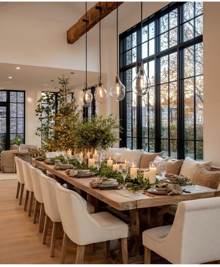 Elegant holiday dining room with rustic wooden table, white chairs, and candlelit centerpiece under pendant lights and large windows.