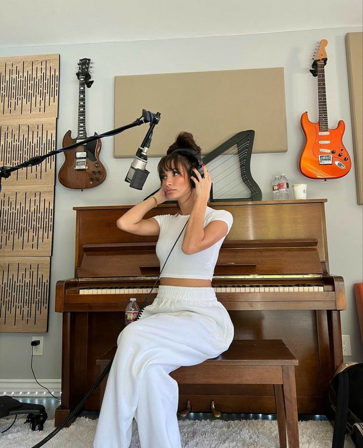 Young woman in a music studio, wearing headphones and sitting at a piano, surrounded by guitars and acoustic panels.