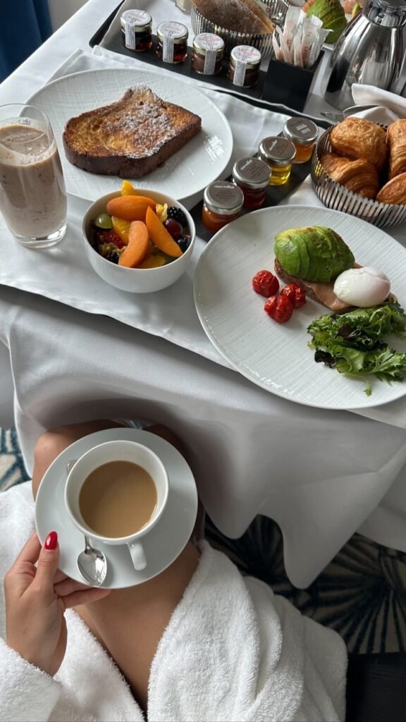 Luxurious breakfast spread with avocado toast, assorted fruits, pastries, coffee, and juice on a white tablecloth.