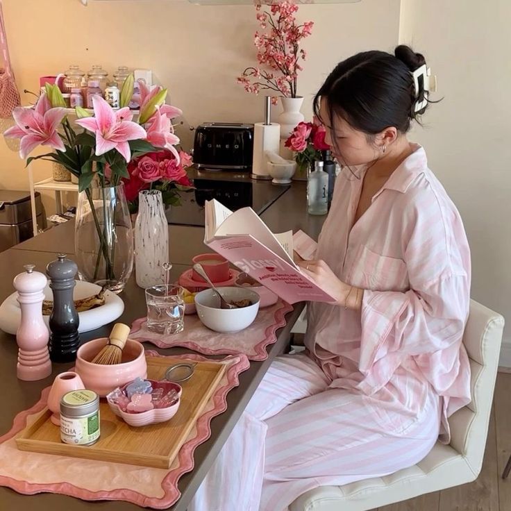 c85246a0-d765-412a-adab-6505a42c9e72 - Handy Home Woman enjoying morning tea and reading at a cozy kitchen table with flowers and breakfast items, wearing striped pajamas.