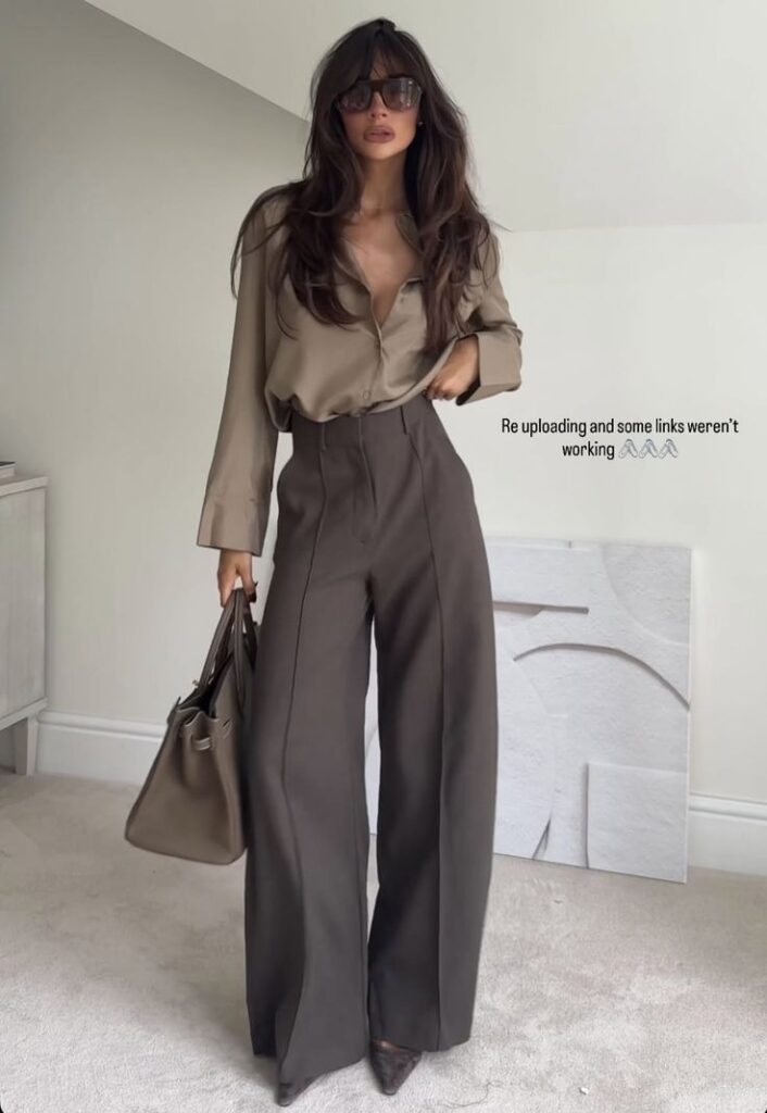 Woman in stylish brown outfit with sunglasses, wide-leg pants, and a handbag, standing in a modern room. Fashionable look.