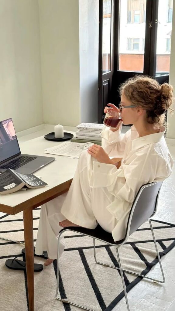 Young woman in white attire drinking coffee while working on a laptop at home office; books and candle on the desk. Cozy workspace.