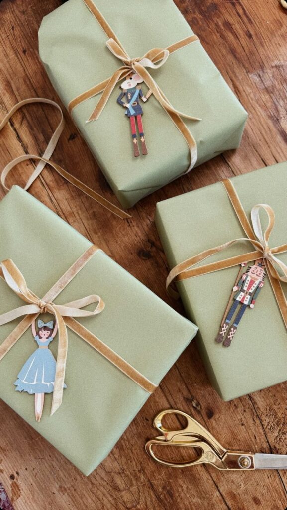 Green gift boxes with gold ribbons and paper doll tags on a wooden table, next to gold scissors. Holiday wrapping.