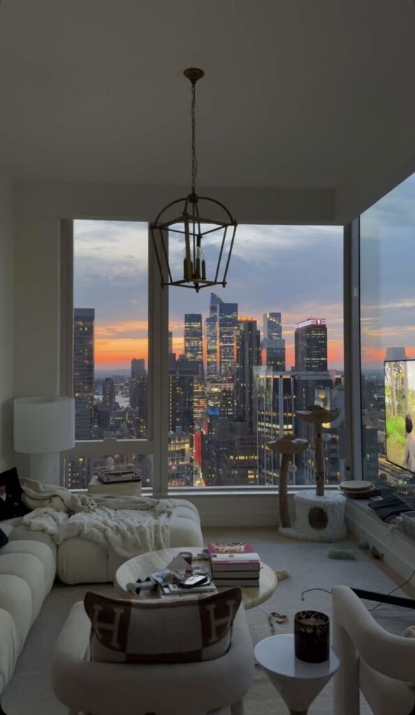 Modern living room with large windows showcasing a vibrant city skyline at sunset. Cozy interior with plush seating and decor.