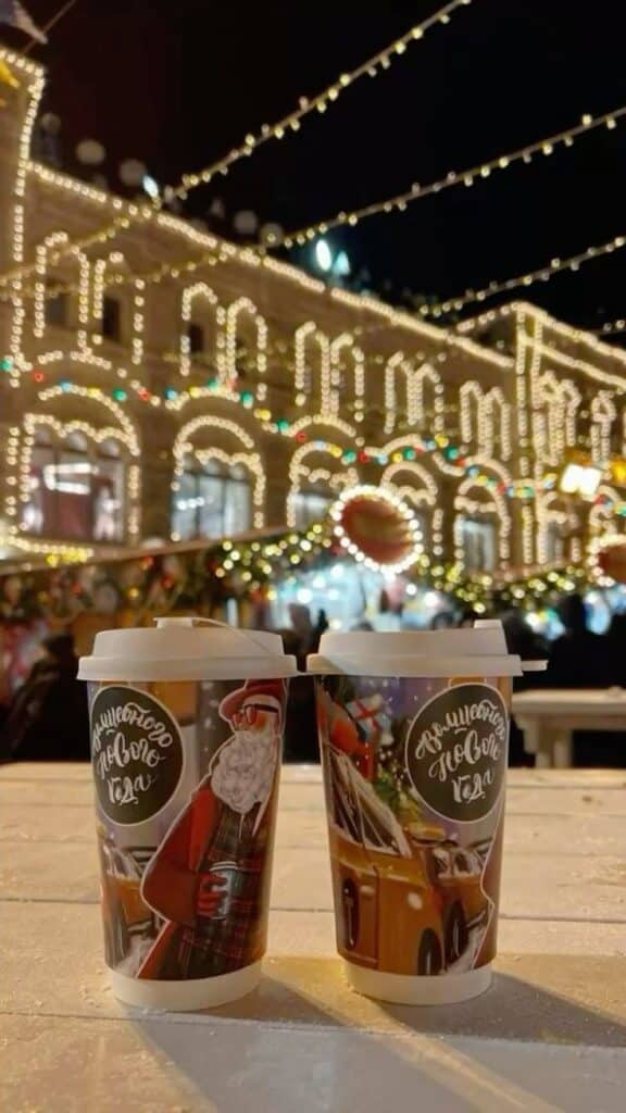 christmas winter hotdrinks cozy - Handy Home Festive cups with Christmas designs in front of illuminated building and decorations at night. Cozy holiday atmosphere.