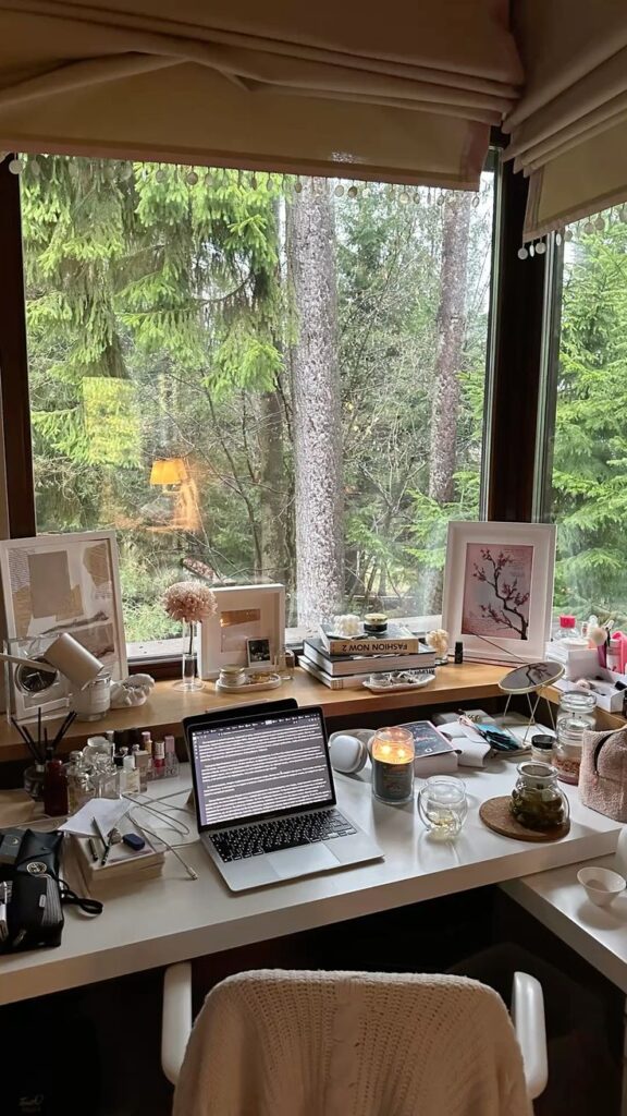 Cozy home office with a laptop, candle, and books by a large window overlooking lush green trees. Perfect workspace inspiration.