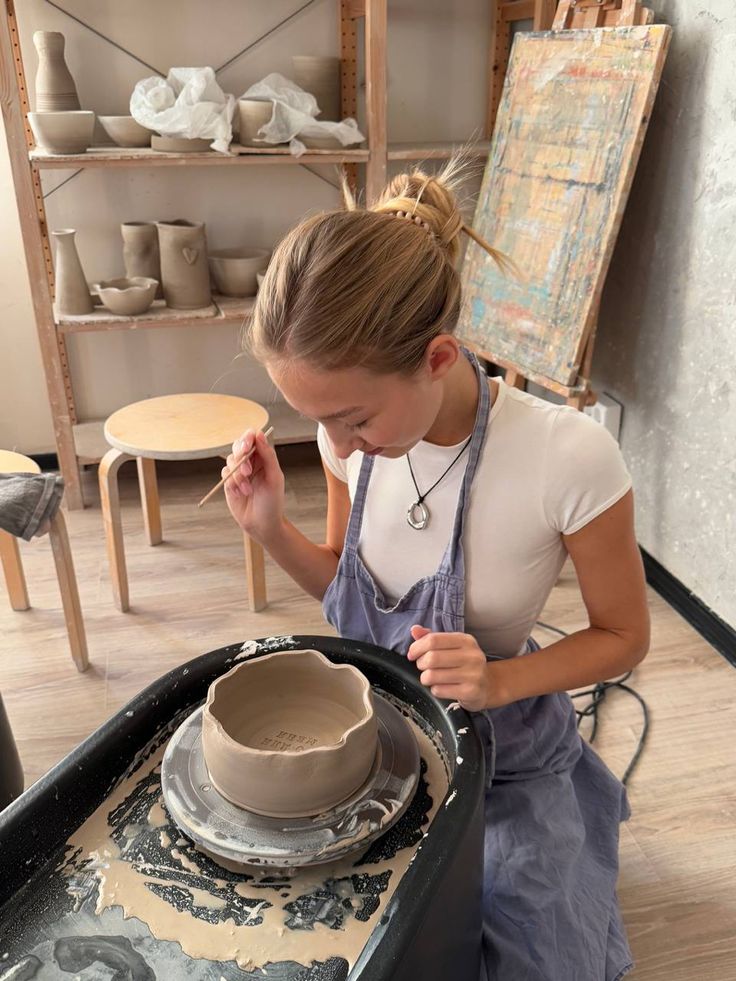 Young woman shaping pottery on a wheel in an art studio, surrounded by clay items and tools.