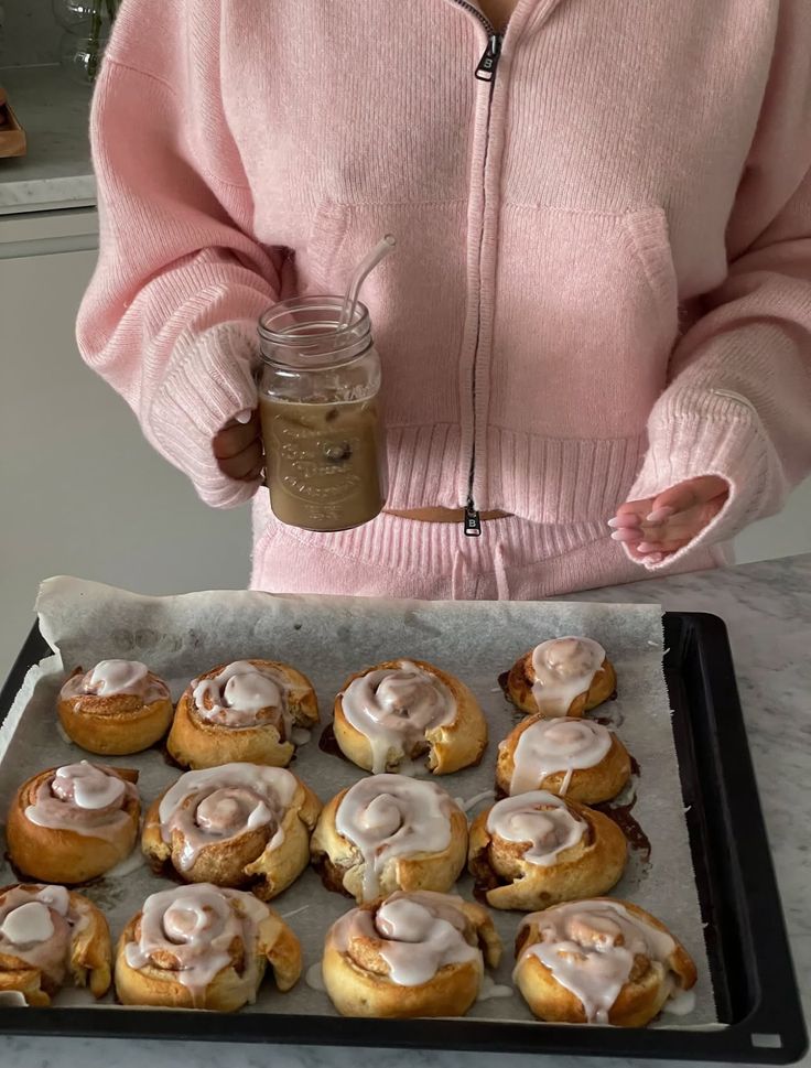 Person in pink sweater holds iced coffee next to a tray of freshly baked cinnamon rolls with icing. Cozy and delicious treat.