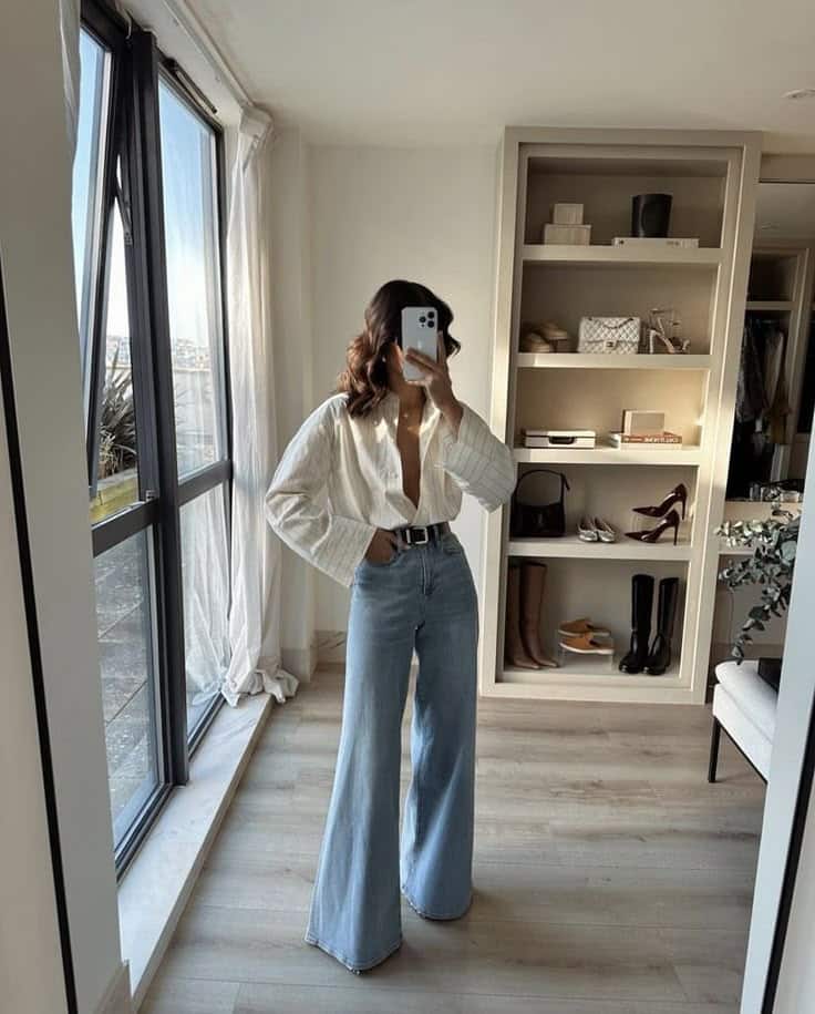 Woman in a white blouse and flared jeans taking a selfie in a modern, stylish room with a shelf of shoes and accessories.