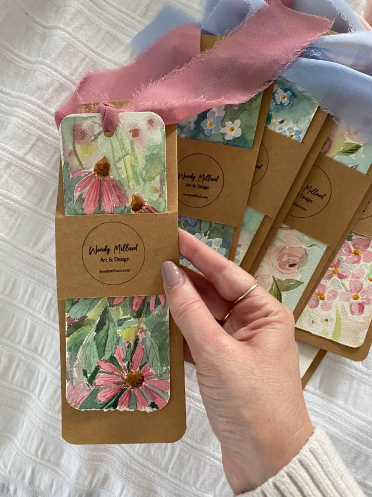 Hand holding floral watercolor bookmarks with pink and blue ribbons on white fabric background.