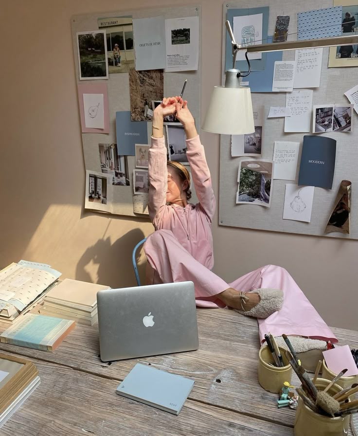 Woman in pink lounging at a desk with a laptop and mood boards in a cozy workspace.