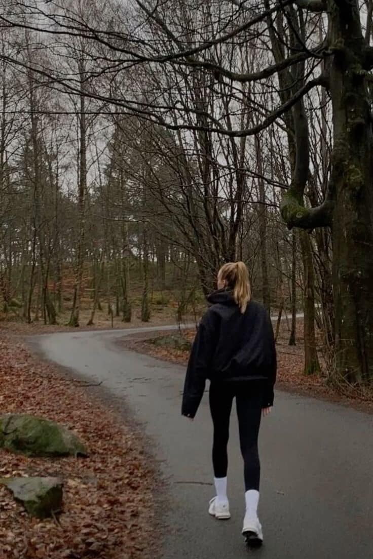 Person walking on a winding forest path during autumn, surrounded by leafless trees and fallen leaves.