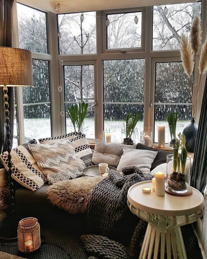 eaf363f5-00dc-4c54-a393-6a4b2b54afd1 - Handy Home Cozy corner by large windows with snow falling outside, featuring pillows, blankets, and candles on a wintry day.