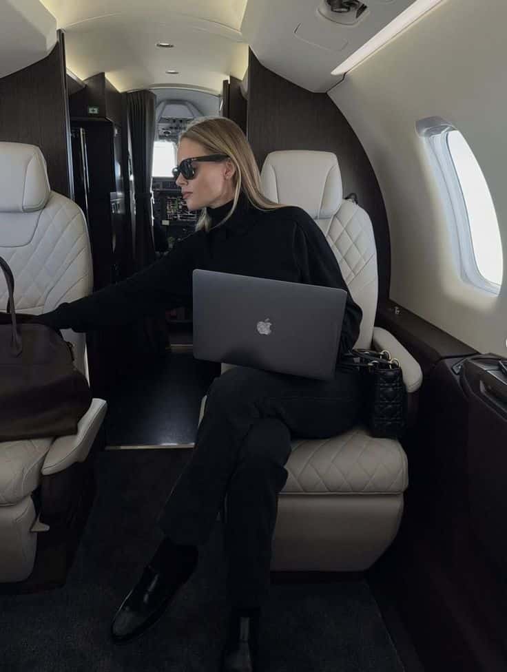 fa5c5299-36d8-45e7-8344-c73f0456ea33 - Handy Home Person with laptop on private jet, wearing sunglasses and black outfit, luxurious travel setting.