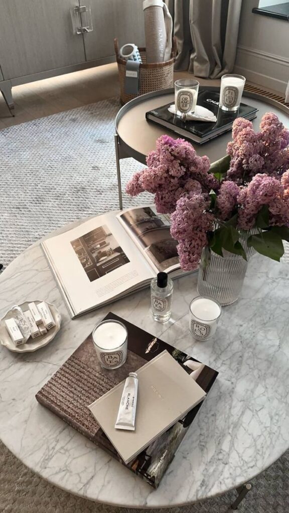 fgsnbtdzfgb - Handy Home Elegant marble table with lilacs, decor books, candles, and skincare products in a cozy living room.