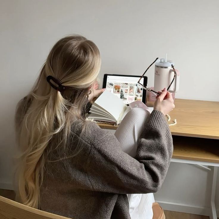 fzgnbzdn - Handy Home Woman reads a book at a desk, holds eyeglasses, with a tablet and drink nearby, cozy indoor scene.