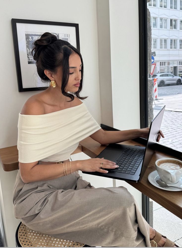 gfb xd - Handy Home Woman working on laptop in a cafe with coffee, wearing off-shoulder top and gold jewelry. Urban view outside the window.