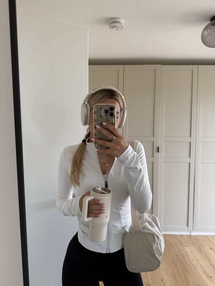 gym fit - Handy Home Person in a white jacket taking a mirror selfie, wearing headphones, with a travel mug and bag in hand, indoors.
