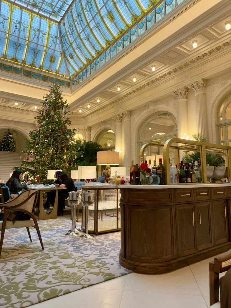 Elegant hotel lobby decorated for Christmas with a grand tree, plush seating, and a stunning glass ceiling, creating a festive ambiance.